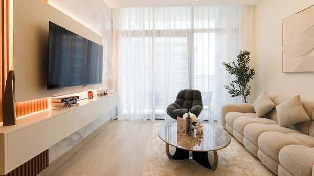 Una sala de estar moderna con un televisor de pantalla plana, un sofá, una mesa de centro y un ventanal con cortinas ligeras.