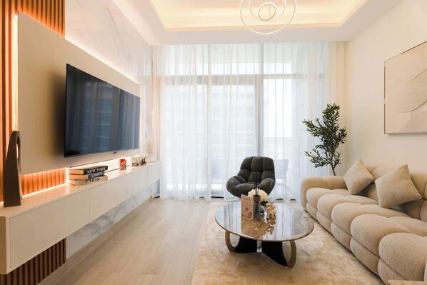 A modern living room with a flat-screen TV, a sofa, a coffee table, and a large window with sheer curtains. - Rentico l Luxury 1BR Pool View l Azizi Riviera (Dubai)