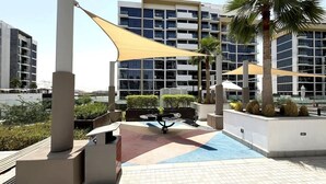 Un área de piscina en la azotea con una sombrilla, palmeras y un edificio moderno al fondo.