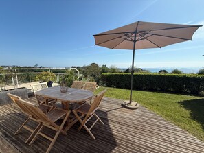 Eine Holzterrasse mit rundem Tisch und Stühlen, einem großen Sonnenschirm und Blick auf das Meer.