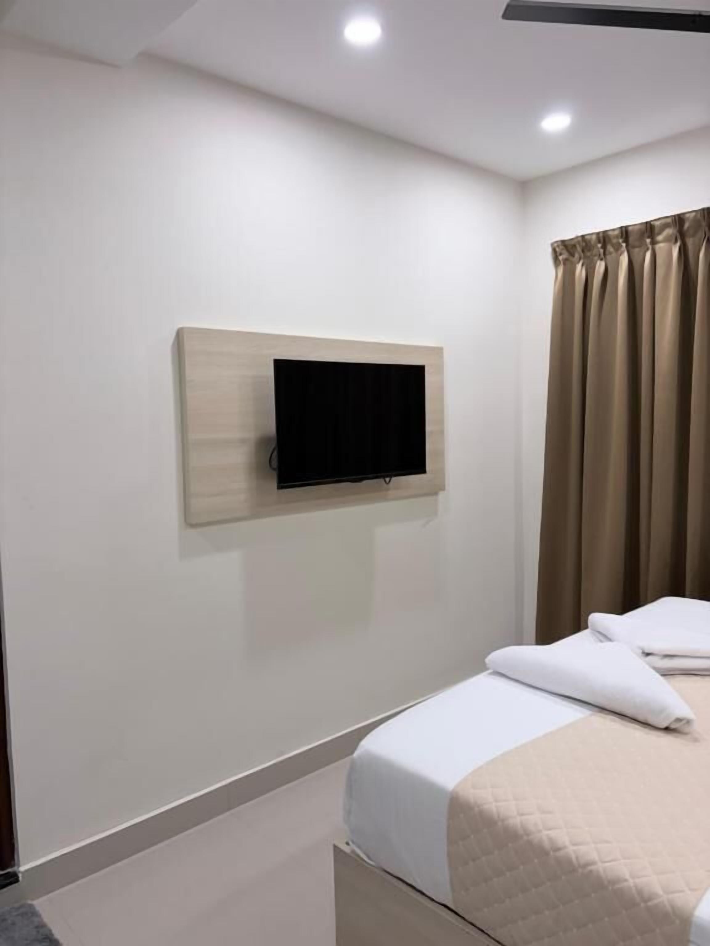 Una habitación de hotel con cama, televisor de pantalla plana montado, ventana con cortinas e iluminación en el techo.