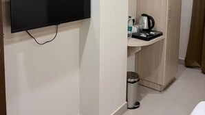 Una habitación con una televisión de pantalla plana en la pared, un armario de madera, un lavamanos pequeño con una tetera y un cubo de basura.