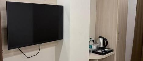 Une chambre dotée d’un téléviseur à écran plat fixé au mur, d’une armoire en bois, d’un petit lavabo avec une bouilloire et d’une poubelle.