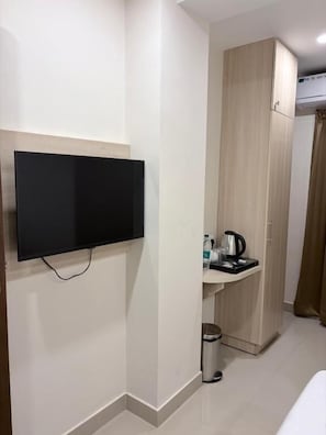 Een kamer met een wandgemonteerde flatscreen-tv, een houten garderobe, een klein wastafeltje met waterkoker en een prullenbak.