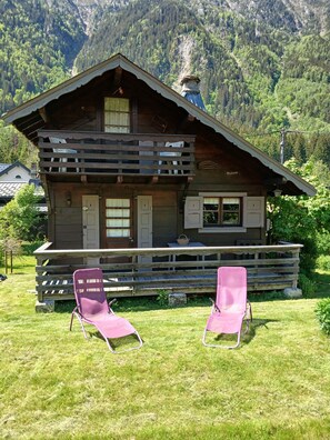 A wooden chalet with a balcony, two pink chairs, and a mountain backdrop.