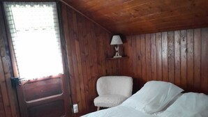 Una habitación con paneles de madera, una cama, una silla, una lámpara en un estante y un ventanal.