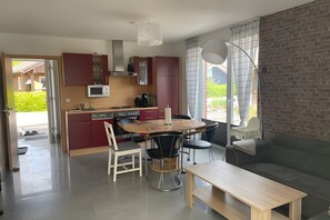 A modern kitchen with a dining area, a sofa, and a view of greenery outside.