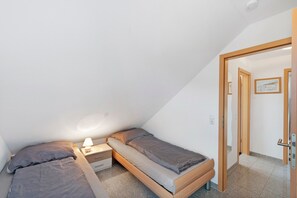 Ein kleines Zimmer mit einem Einzelbett, einem hölzernen Bettgestell, einem Nachttischchen mit Lampe und einer Tür zu einem weiteren Raum.