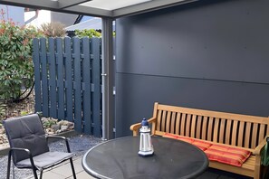 Een terras met een ronde tafel, een houten bankje, een klein decoratief vuurtoren en een blauwe omheining.