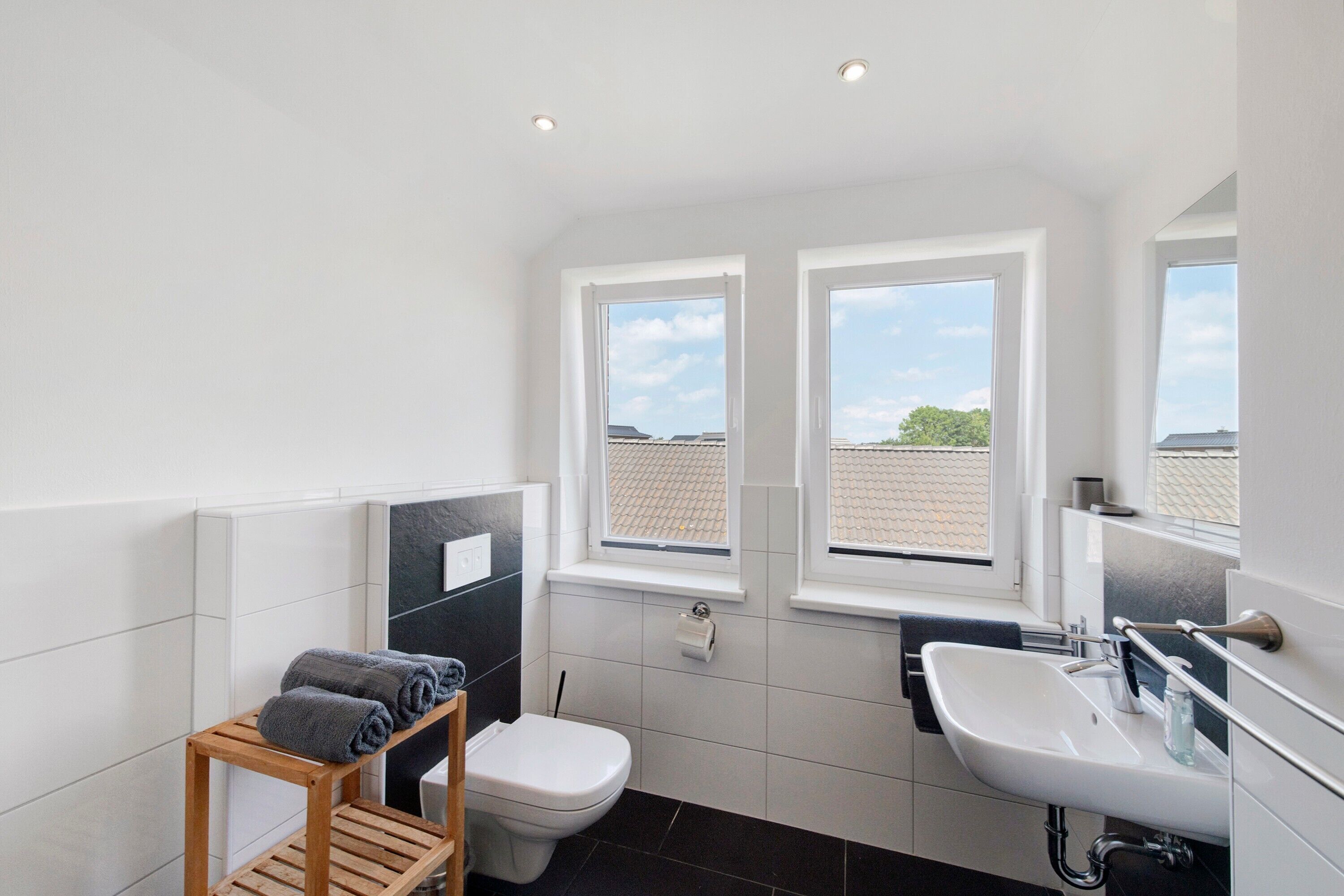 Ein modernes Badezimmer mit Toilette, Waschbecken und einem hölzernen Ablagebereich mit Handtüchern. Drei Fenster sorgen für natürliches Licht.
