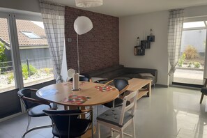 Een moderne woonkamer met een ronde houten tafel, zwart-witte stoelen, een grijze bank en een bakstenen muur.