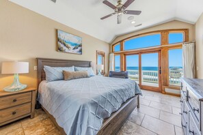 A bedroom with a bed, nightstand, lamp, ceiling fan, and a large window with a view of the ocean.