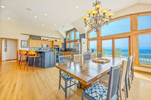 A spacious kitchen with a large island, wooden dining table, and chairs, and a view of the ocean through large windows. - Oceanfront, Dog Friendly Home in Hatteras with Pool, Hot Tub, Game Room (Hatteras)