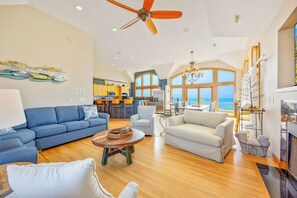 A spacious living area with a ceiling fan, a kitchen, and large windows offering a view of the ocean. - Oceanfront, Dog Friendly Home in Hatteras with Pool, Hot Tub, Game Room (Hatteras)