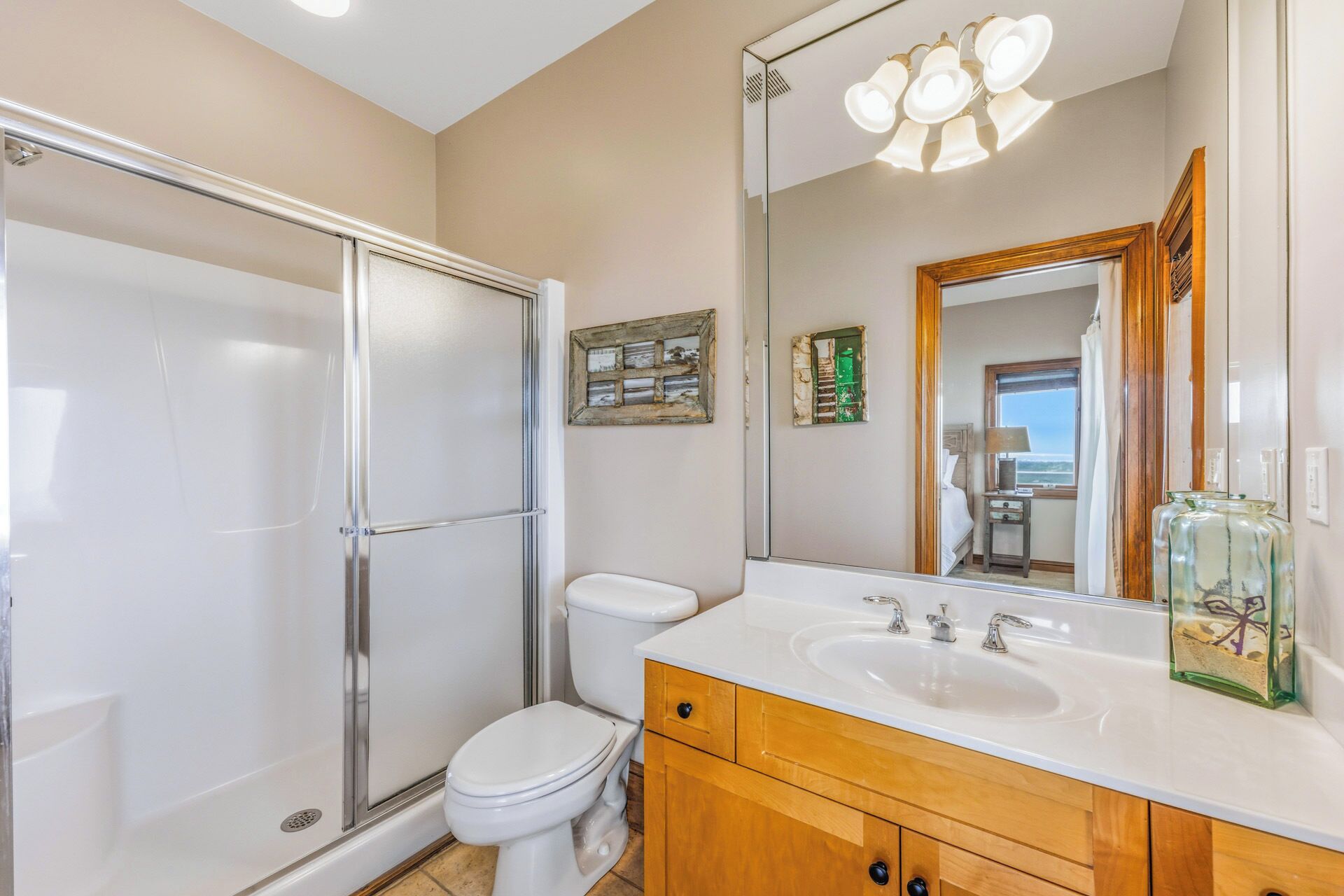 A bathroom with a glass shower enclosure, a toilet, a sink with a mirror, and a view of a room with a bed and window.