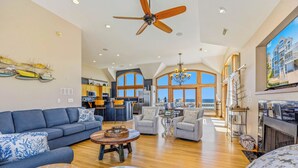 A spacious living area with a large sofa, a coffee table, and a fireplace. There is a kitchen in the background with a view of the ocean.
