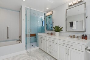 Un baño moderno con cabina de ducha de vidrio, un tocador blanco con espejo y una planta en maceta.