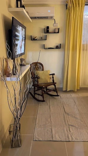 Una sala de estar con televisor, una mecedora, un tapete estampado y objetos decorativos en estantes.