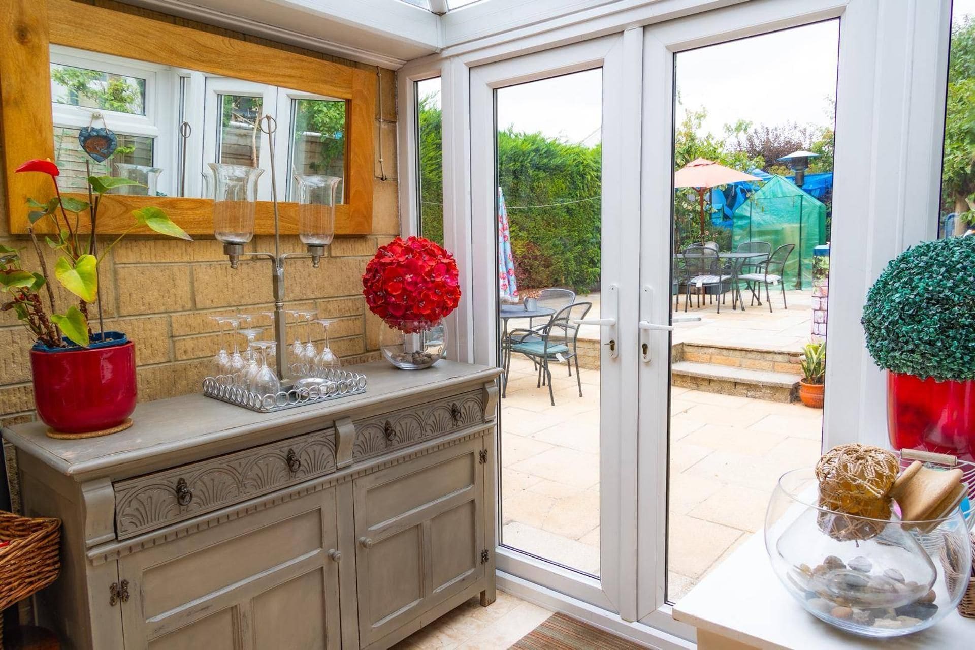 A conservatory with a glass door leading to an outdoor patio area. Inside, there is a wooden cabinet with glassware and a potted plant.