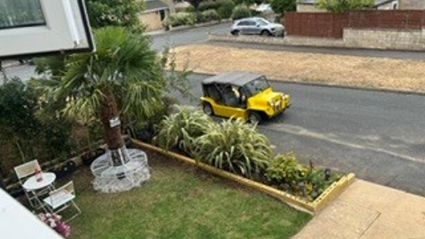 A residential area with a lawn, a small garden bed, and a parked yellow vehicle. - House Sleeps 6 With Enclosed Garden (Cirencester)