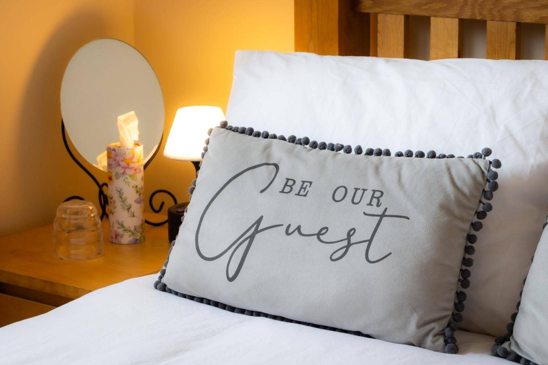 Un lit avec un oreiller sur lequel est brodé le message « SOYEZ NOTRE INVITÉ » et une table de chevet avec une lampe et un vase décoré de motifs floraux.