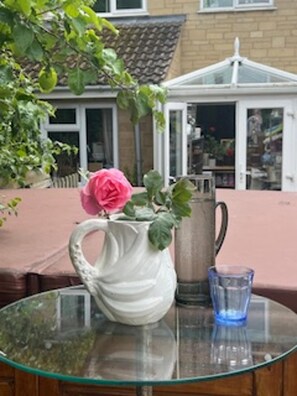 A glass table with a white pitcher and a pink flower, a blue glass, and a brick wall in the background.