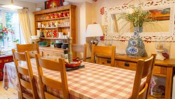 A dining area with a checkered tablecloth, wooden chairs, a shelf with dishes, and a vase with flowers on the wall.