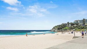 Uma praia com céu claro, ondas do oceano e um edifício com vários andares e varandas.