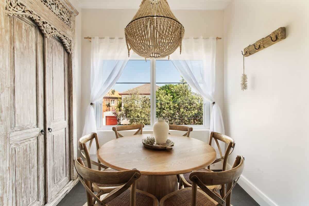 Un comedor con una mesa redonda de madera, seis sillas, una lámpara colgante de fibras tejidas y vistas amplias a través de grandes ventanales.