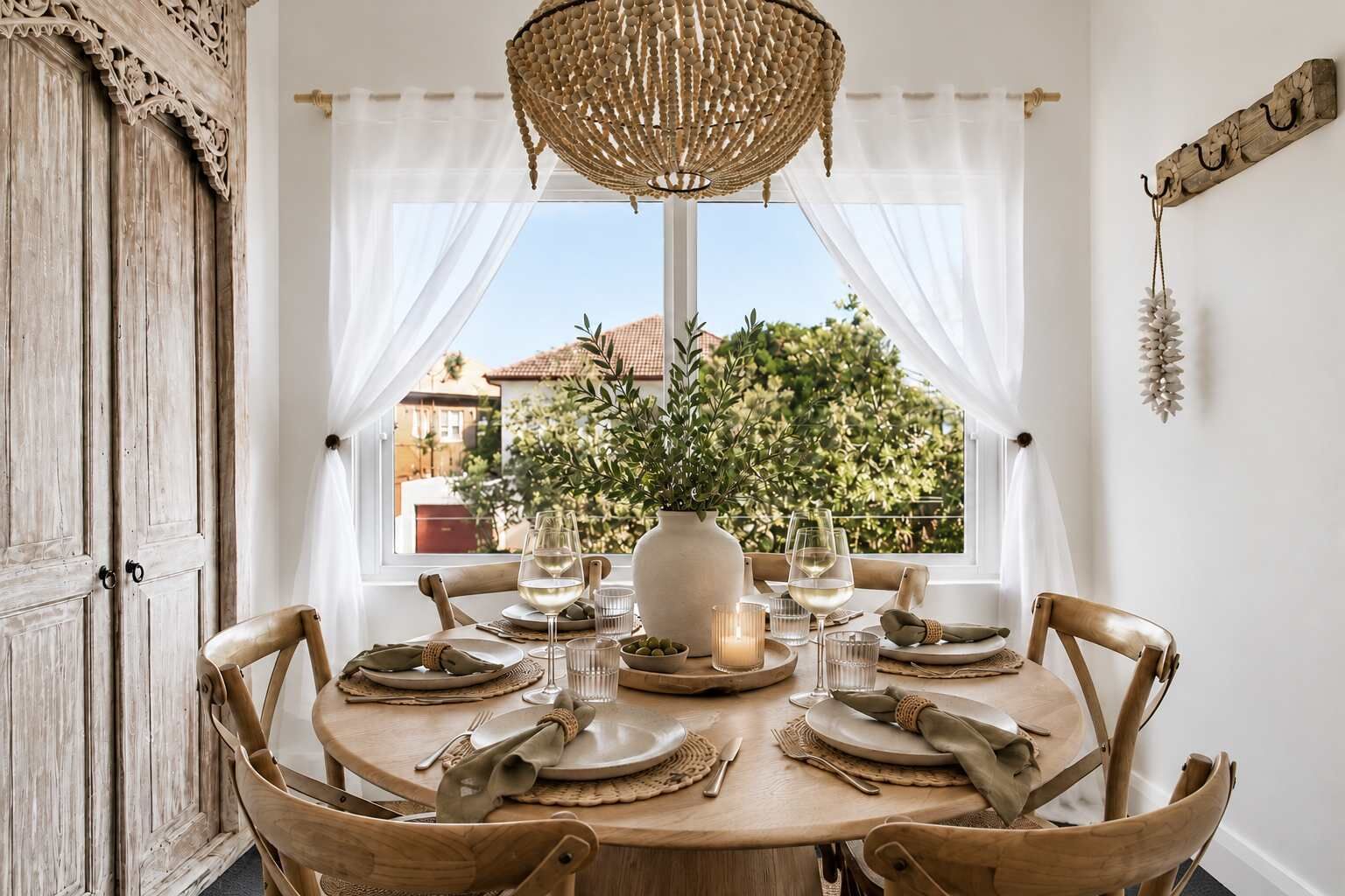 A round dining table set for four with a rustic wooden finish, white tablecloth, and a woven chandelier above.