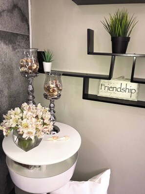 Uma toucador de banheiro com pia branca, dois castiçais de vidro com conchas, um vaso de flores brancas, uma planta em vaso e uma prateleira com a placa 'Friendship'.
