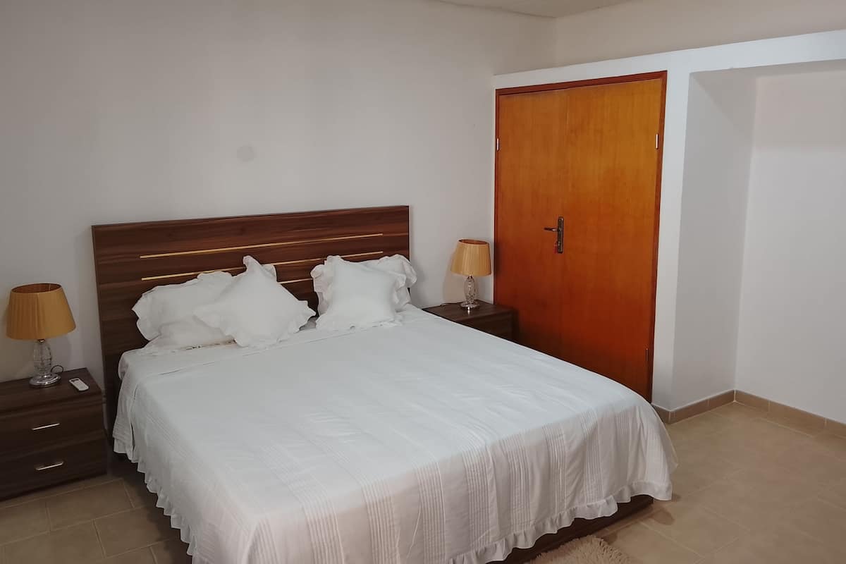 Un dormitorio con cabecera de madera, ropa de cama blanca, dos lámparas de noche, un armario de madera y suelo de baldosas.