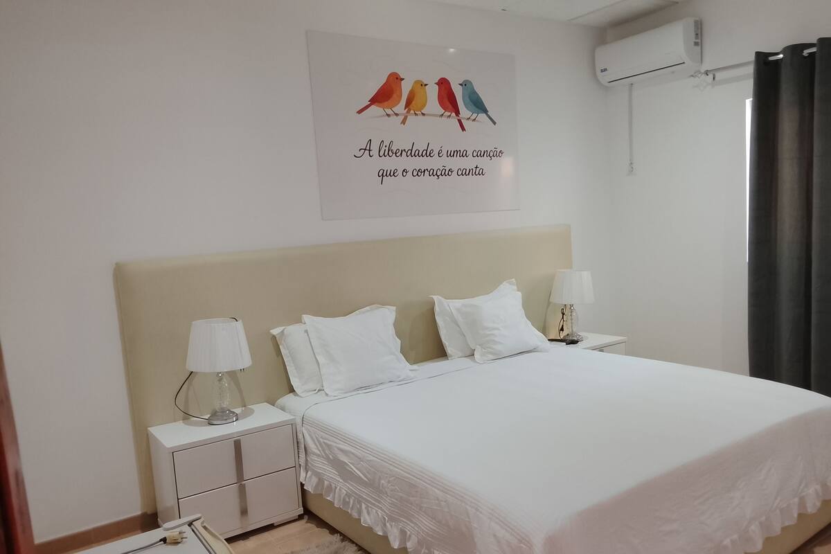 Habitación de hotel con cama, dos mesitas de noche, lámpara, un adorno en la pared y un equipo de aire acondicionado.