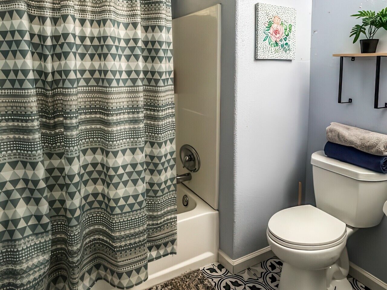 A bathroom with a patterned shower curtain, a toilet, a bathtub, a wall-mounted shelf with a plant, and a framed artwork.