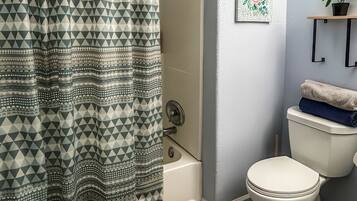 A bathroom with a patterned shower curtain, a toilet, a bathtub, a wall-mounted shelf with a plant, and a framed artwork.