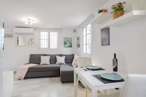A modern living room with a grey sofa, a dining table with a wine bottle, and a white wall with artwork. - Sunny Corner Apt in Puebla Aida Golf (Mijas)