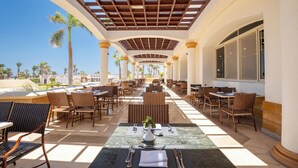 Un espacio de comedor al aire libre con mesas y sillas, una pérgola y palmeras al fondo.