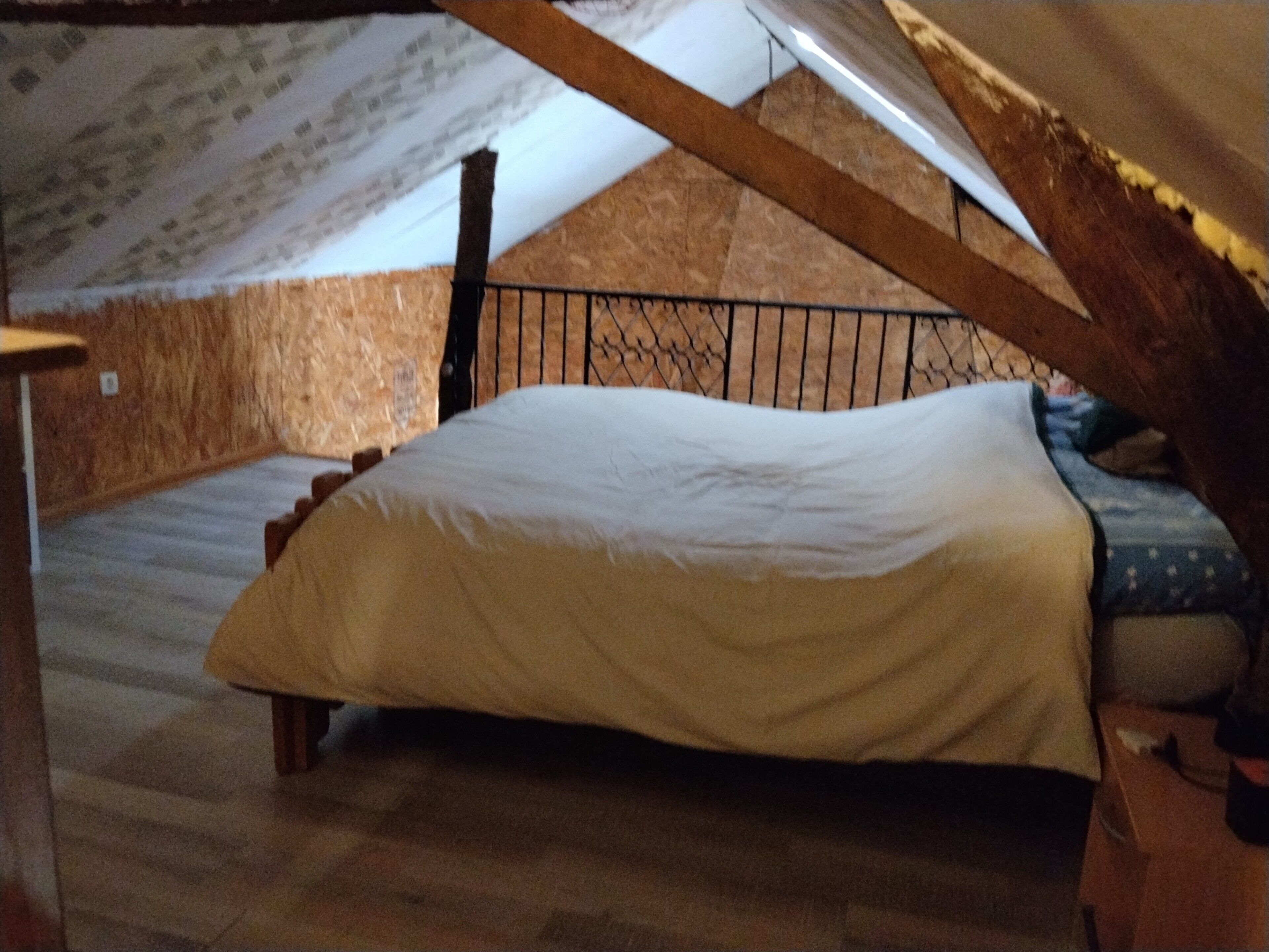 Ein Zimmer mit schrägem Dach, einem Bett mit weißer Matratze, Holzboden und einem Metallgeländer.