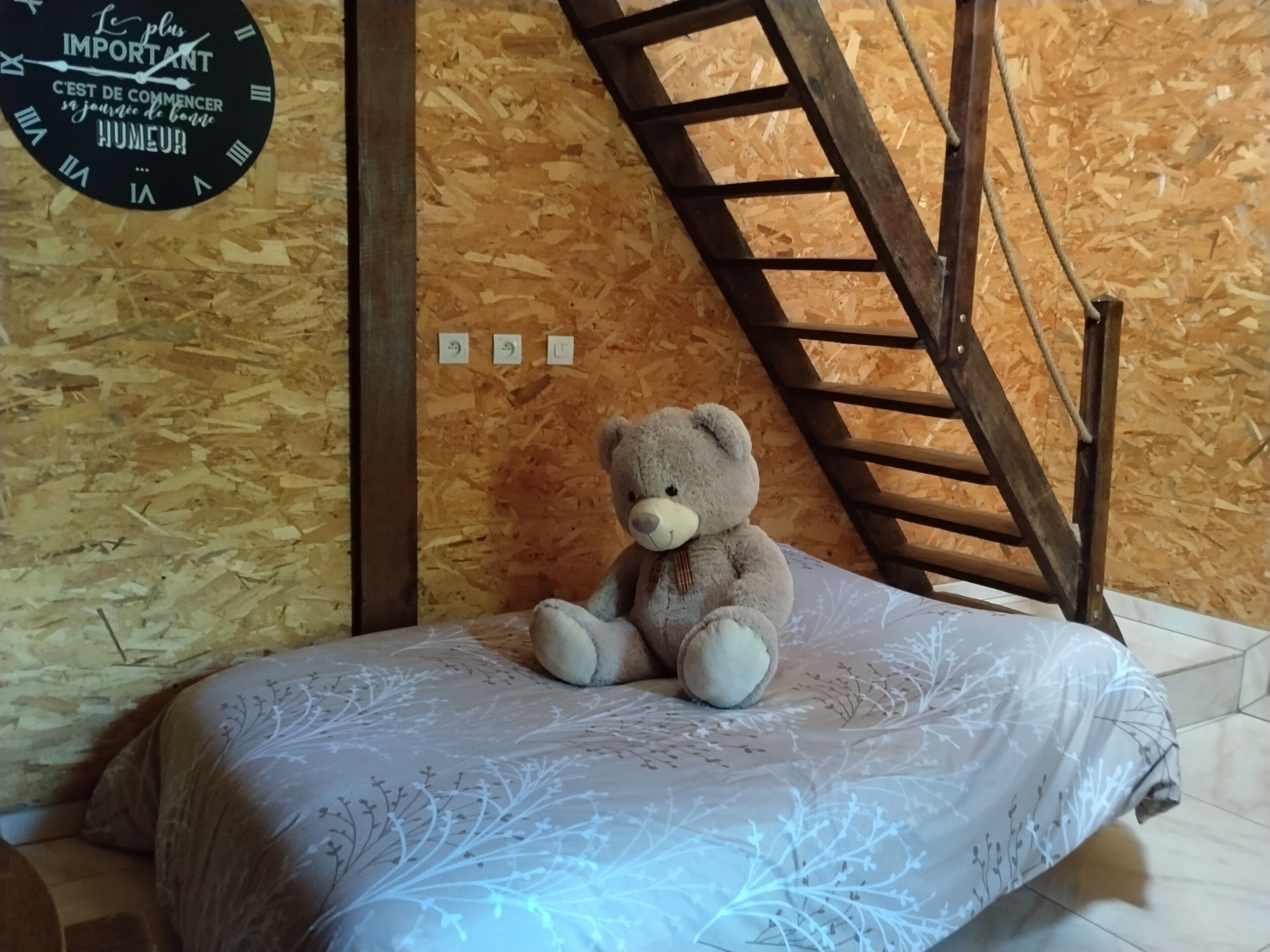 Une pièce avec un plafond en pente en bois, un lit recouvert d’une courtepointe à motifs, un ours en peluche sur le lit, une horloge murale et une échelle en bois.