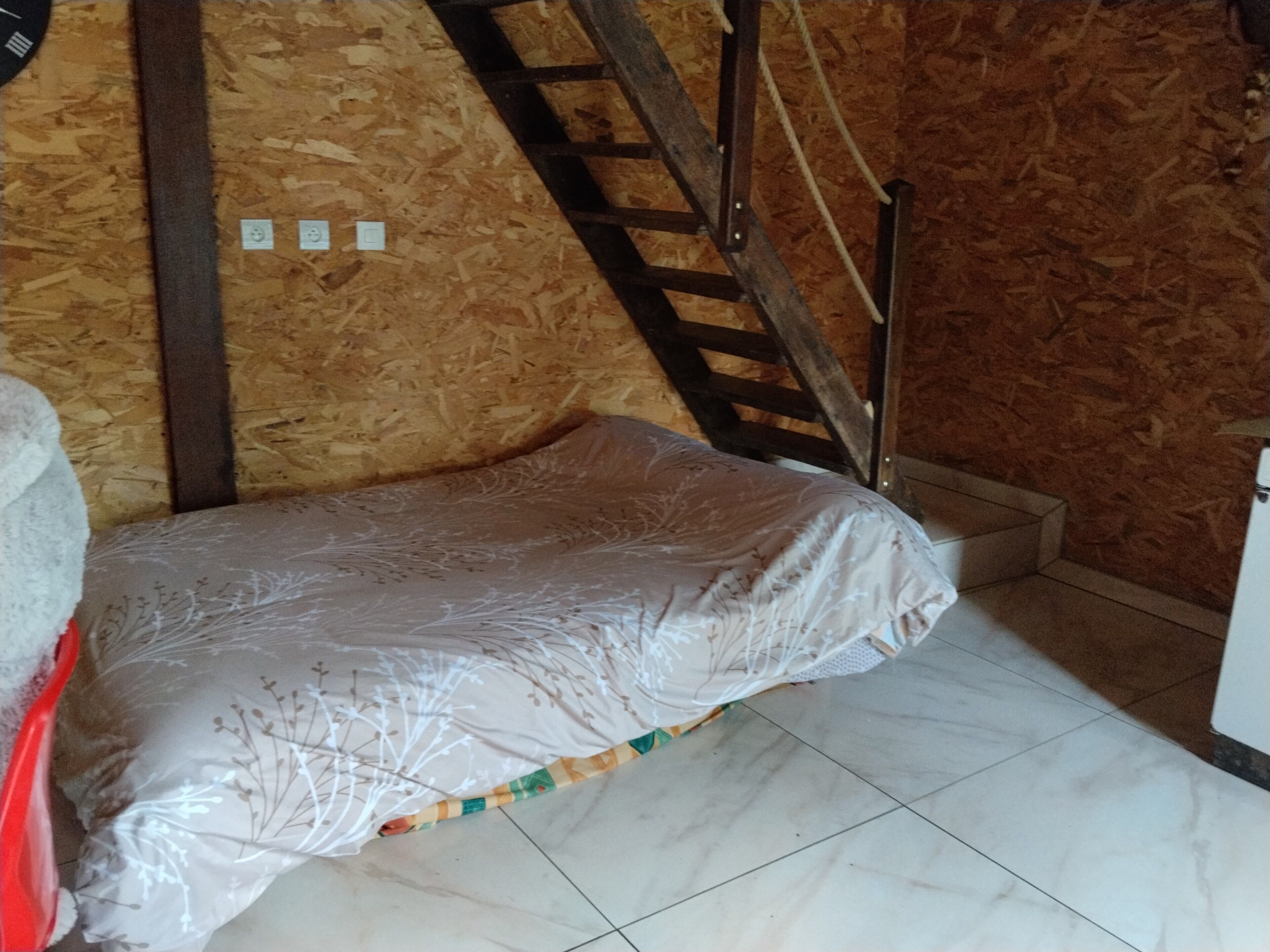 Pequeña habitación con cama, una escalera de madera y suelo de baldosas.
