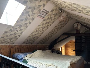 Una habitación con techo inclinado, papel tapiz estampado, una cama con manta blanca, un reloj y una puerta de madera.