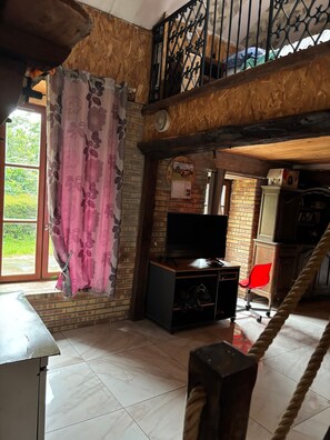 Una habitación con pared de ladrillo, techo de madera, un televisor sobre un mueble de madera, una silla roja y una escalera.