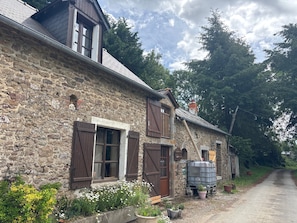 Un bâtiment en pierre avec des volets en bois, un réservoir d’eau en métal et une allée gravillonnée.