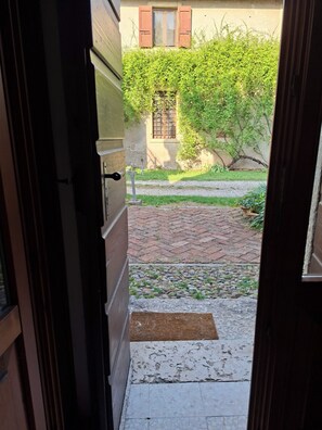 Eine offene Tür, die zu einem Innenhof mit einem Ziegelweg, einer Fußmatte und einem Gebäude mit Fenster und Rollläden führt.