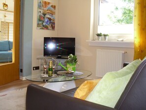 A living room with a glass coffee table, a flat-screen TV, a sofa, and a window with a view of greenery. - Apartment 'Vacation Home Haus Am Wald' with Wi-Fi (Drolshagen)
