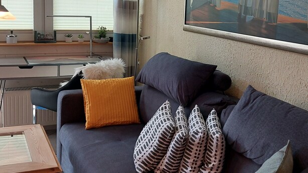 Un salon moderne avec un canapé foncé, des coussins à motifs, une lampe et une grande fenêtre donnant sur un beau paysage.
