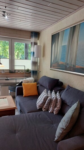 A modern living room with a dark sofa, patterned cushions, a lamp, and a large window with a view. - Apartment "Relax in The House By The Forest" with Wi-Fi (Drolshagen)