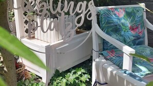 Un espace extérieur aménagé avec des sièges blancs, agrémenté d’une enseigne décorative, entouré de verdure et bordé d’un mur en pierre.