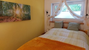 Ein Schlafzimmer mit Bett, einem Fenster mit Vorhängen, einem Bild an der Wand und einem Nachttisch.