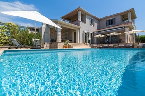 Ein Schwimmbecken mit Liegestühlen und Sonnenschirmen, ein mehrstöckiges Gebäude mit Balkonen und ein klarer, blauer Himmel.